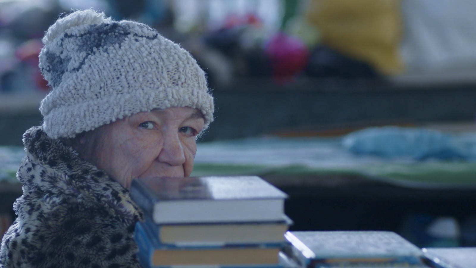 An elderly woman at a market, standing behind a stack of books